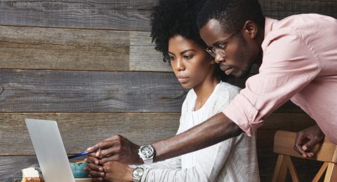 People and education concept. Young African teacher in glasses explaining new material on economics to his beautiful female student, pointing at laptop screen with pen while having lesson at cafe